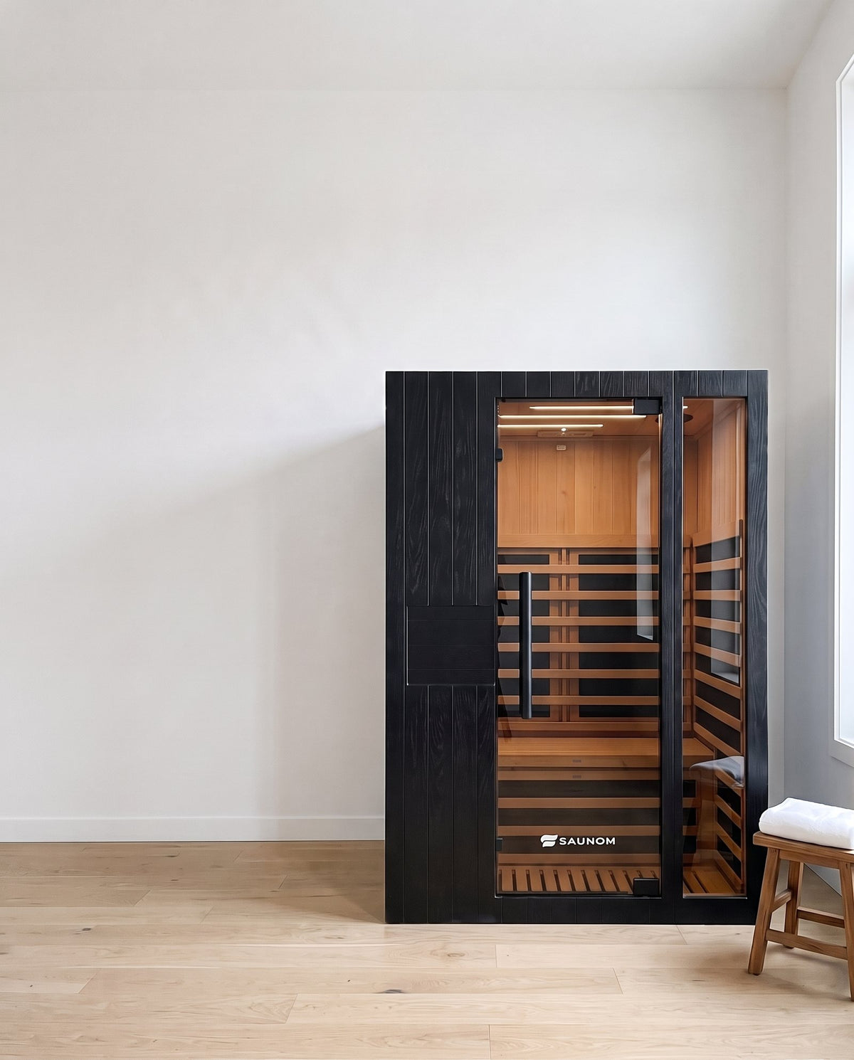 Black Saunom infrared sauna in a calm spare room next to a wooden stool