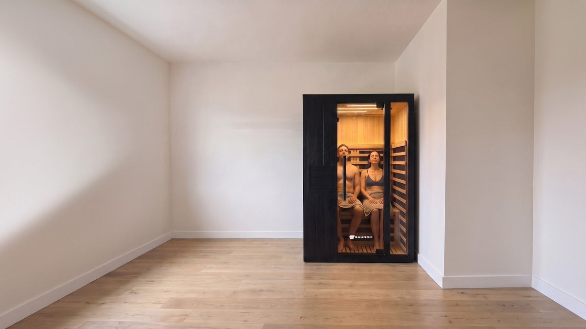 Two adults sitting side by side and leaning back inside a black Saunom infrared sauna, seen through the tinted glass door