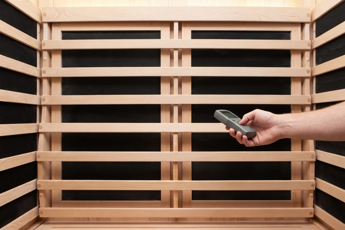 A hand holding a small EMF meter a few inches from a flat heater panel inside a bright infrared sauna cabin.