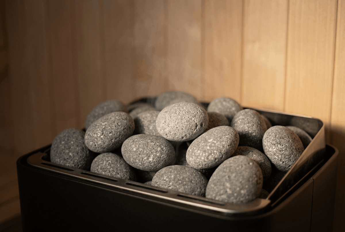A residential electric sauna heater filled with loosely stacked gray volcanic sauna stones with white speckling, in a warm pale-wood Finnish sauna.