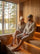 A man and a woman relax on wooden sauna benches inside a wood-paneled sauna with a large window overlooking a forest and lake. The man wears a grey sauna bucket hat and dark swim shorts, and the woman wears a white merino sauna hat and a black swimsuit.