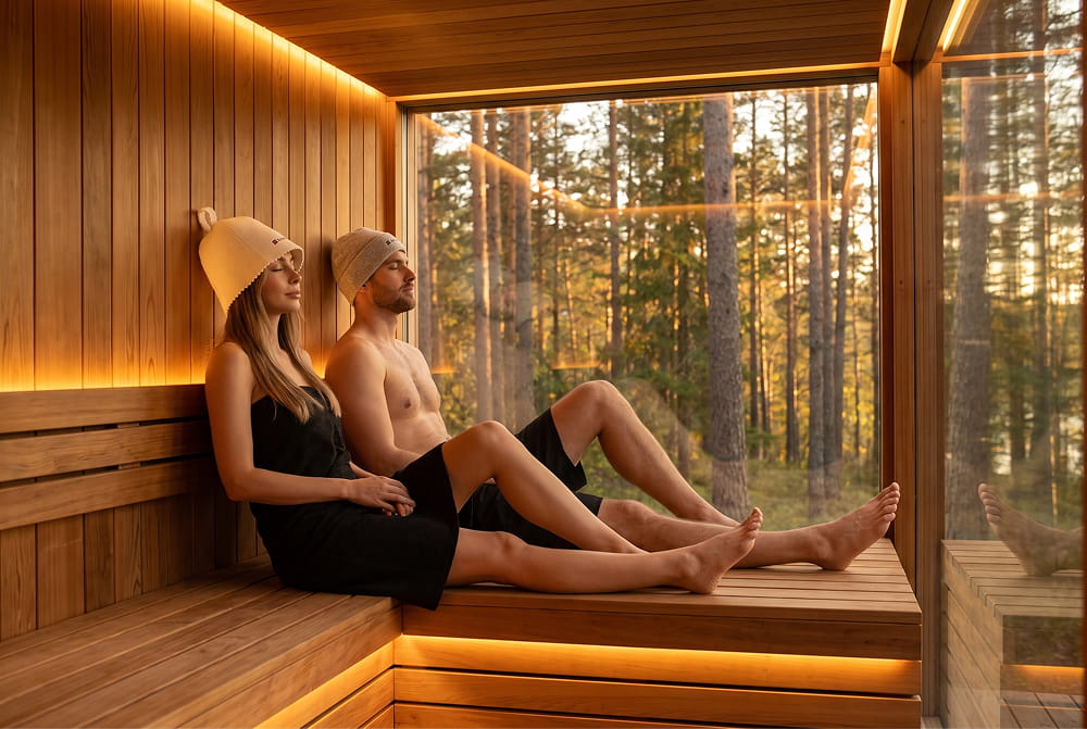 A woman with her long hair tucked under a cream wool sauna hat and a man in a grey wool beanie relaxing in a warm cedar sauna.