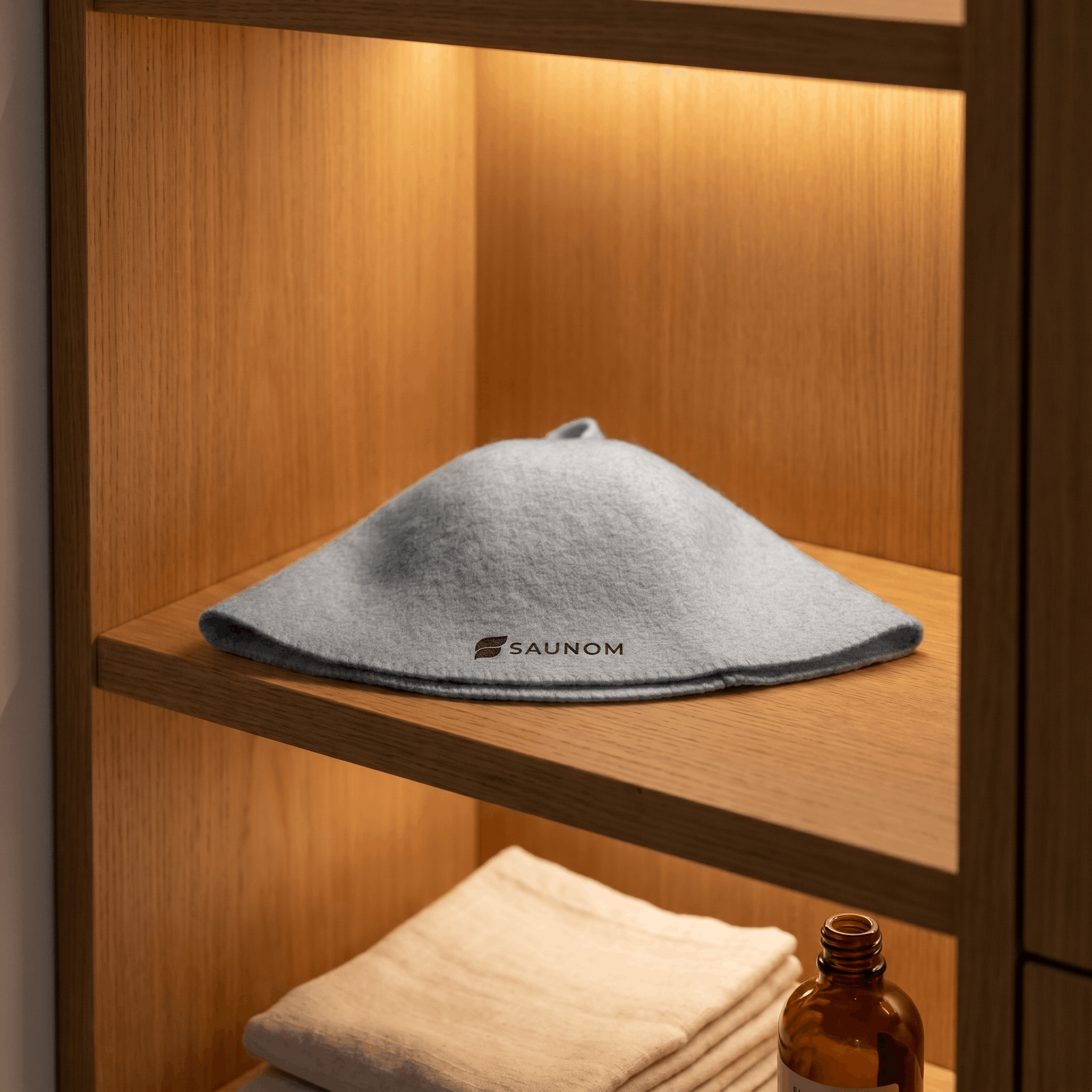 Grey wool sauna hat displayed flat on a wooden shelf with towels and amber bottle