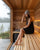 A woman sits barefoot on a wooden sauna bench inside a wood-paneled sauna, wearing a sauna gray beanie and a black dress, with steam rising and a window showing a misty lakeside forest outside.