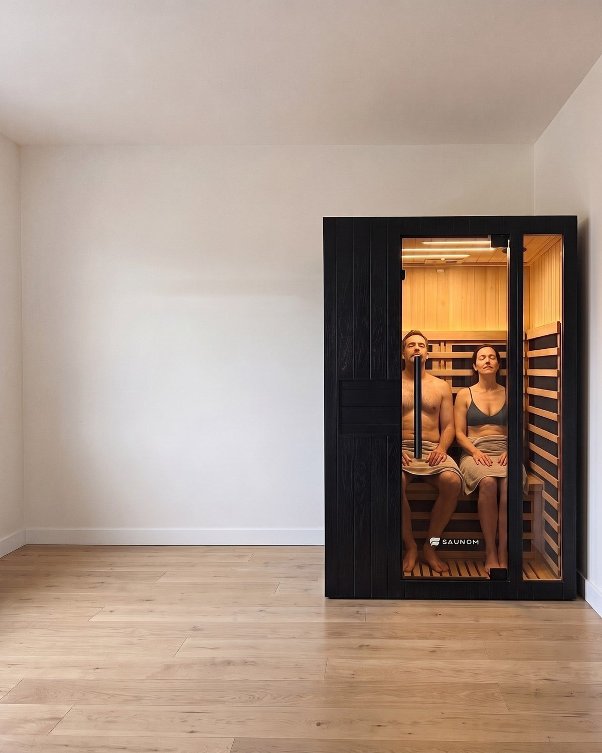 Two adults sitting side by side and leaning back inside a black Saunom infrared sauna, seen through the tinted glass door