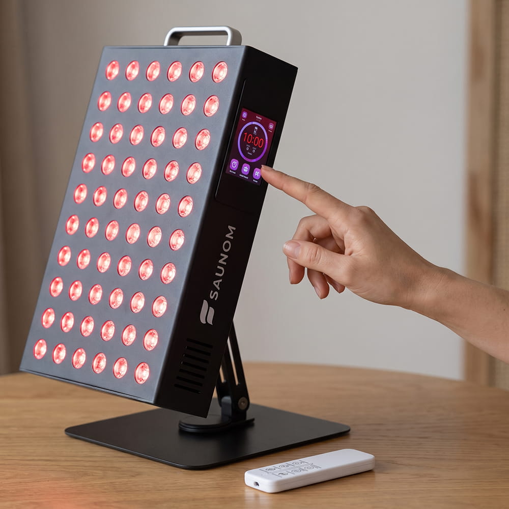 Hand tapping the side touchscreen of the Saunom EW300 Pro red light therapy panel, with the remote beside it