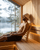 Man relaxing on a curved wooden sauna backrest while wearing a sauna hat inside a lakeside sauna