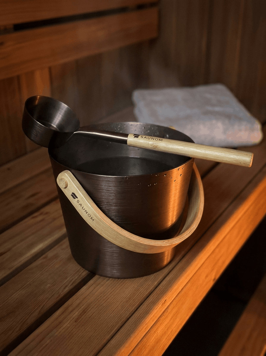 Close-up lifestyle photo of a SAUNOM bamboo-handled sauna bucket with matching ladle resting on a wooden sauna bench