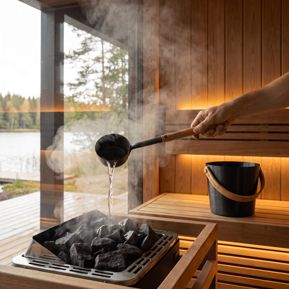 Water poured from a black SAUNOM ladle onto sauna stones to create steam, with the matching bucket behind.