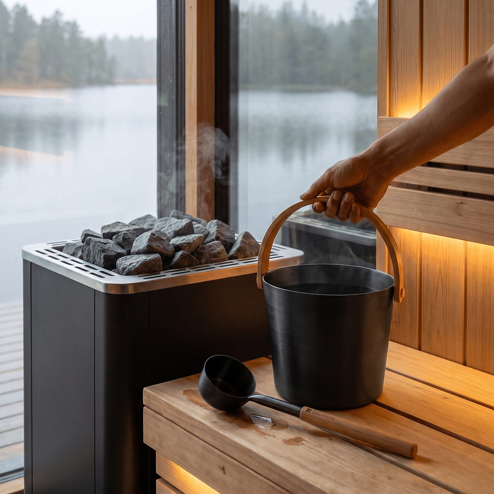 SAUNOM black sauna water bucket held by its bamboo handle, with a matching ladle on the bench beside the heater.