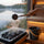 SAUNOM ladle pouring water onto hot sauna stones, with a black water bucket on the bench overlooking a lake.