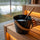 Close-up of a SAUNOM black sauna water bucket filled with water, with a bamboo-handled ladle resting inside.