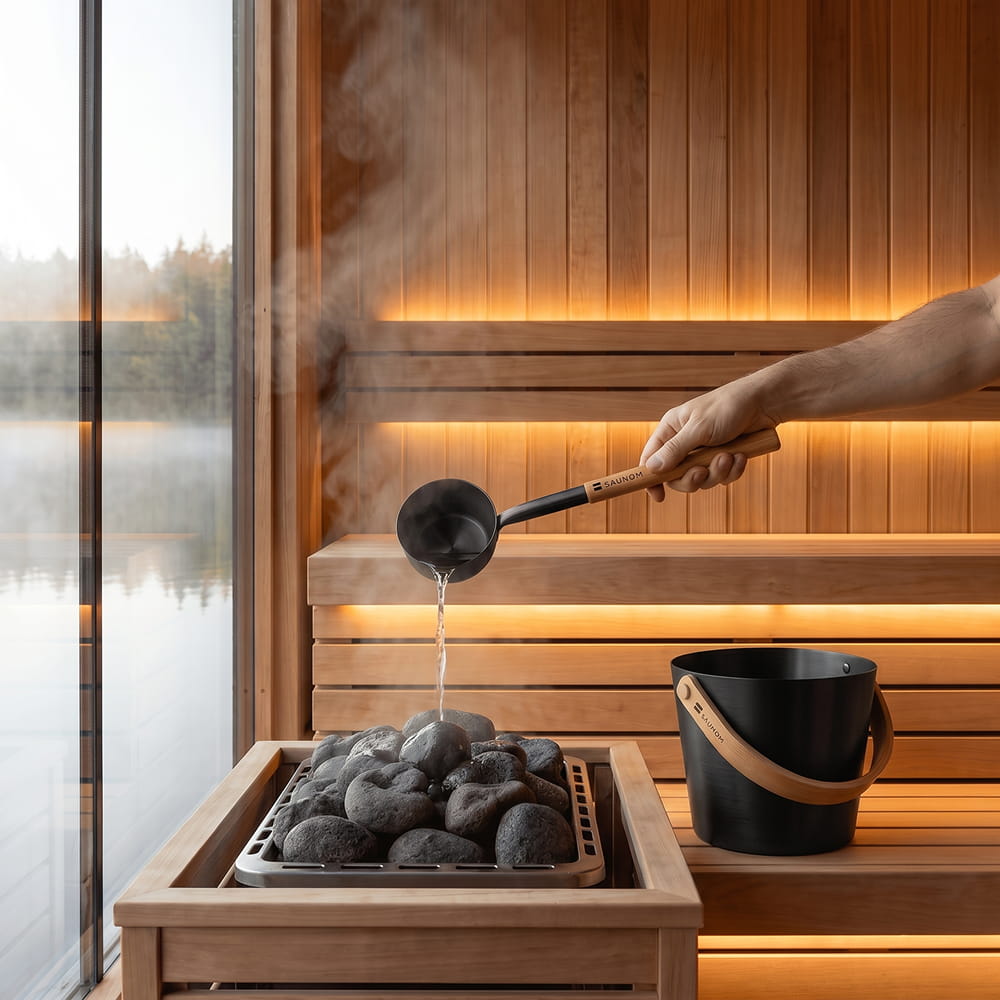SAUNOM black sauna bucket beside a heater as a matching bamboo-handled ladle pours water over the stones.