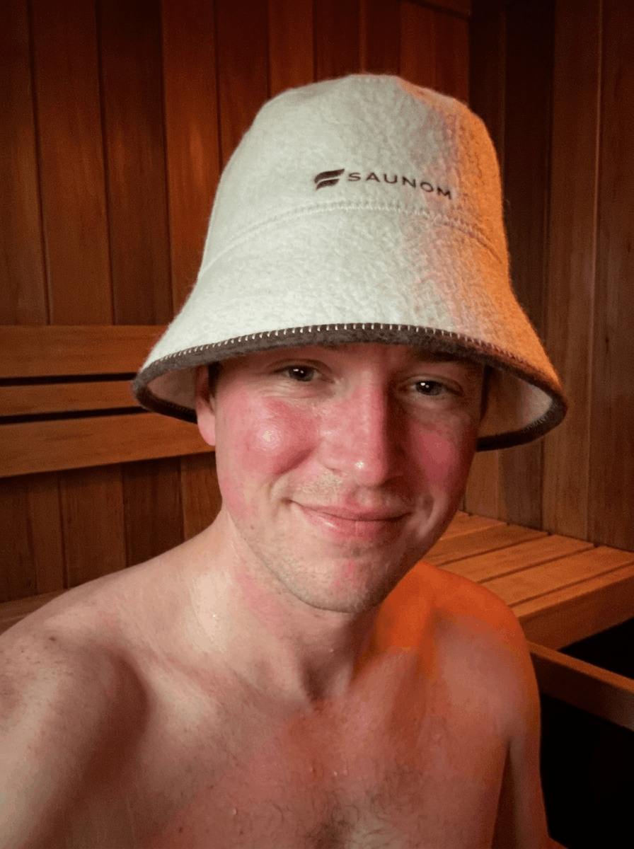 Man smiling inside a wooden sauna while wearing a white bucket-style sauna hat
